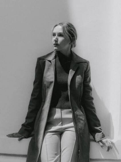 A black-and-white photo shows Rose-Ayling Ellis, wearing a long coat and a sweater, reclining on a wall.