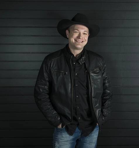Saskatchewan country recording artist, Chris Henderson. Smiling man wearing black cowboy hat, black leather jacket, black shirt and blue jeans.