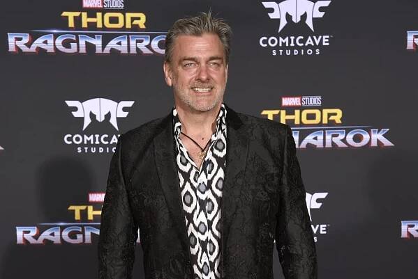 Ray Stevenson in a dark blazer and a patterned dress shirt smiling at a red carpet
