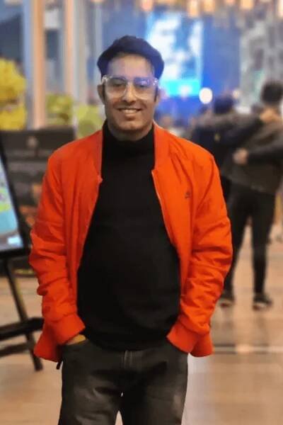 Ein Mann in einer orangefarbenen Jacke und einem schwarzen Poloshirt lächelt direkt in die Kamera. Er steht draußen in einer Art Café. 