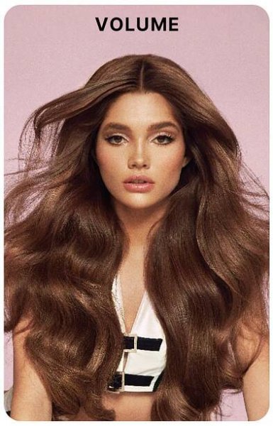 Model with long, full-bodied, voluminous brunette hair styled in loose waves, wearing a white top with black accents against a soft pink background. Bold black text above reads: “VOLUME.”