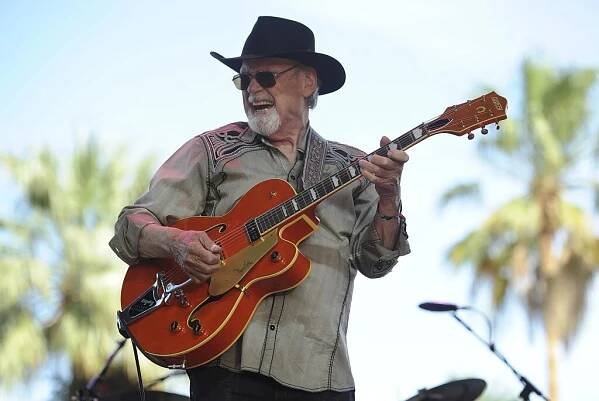 A man in a grey button down shirt and a black, wide-brimmed hat on a stage playing a guitar