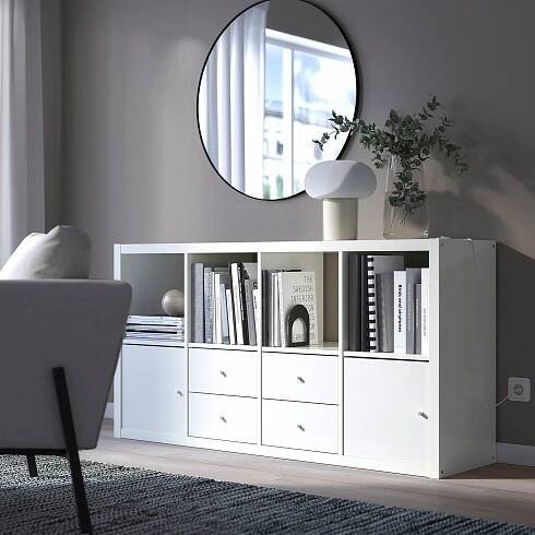 Bílá police IKEA KALLAX s knihami, dekoracemi, rostlinami a lampou. Velké zrcadlo odráží okno. Šedá pohovka, koberec, útulný minimalistický prostor.