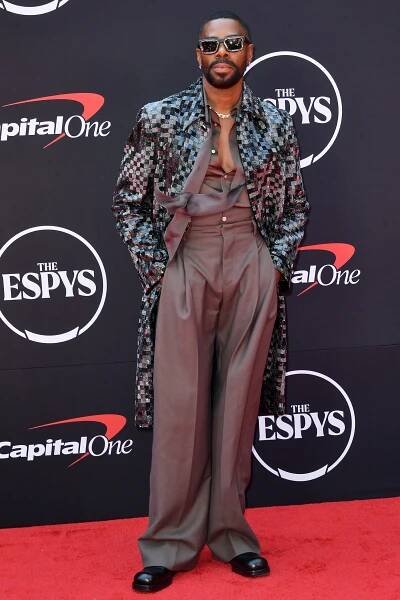 Colman Domingo posing on a red carpet wearing a grey overcoat, unbuttoned top and trousers. He is wearing sunglasses and standing with his hands in his pockets.