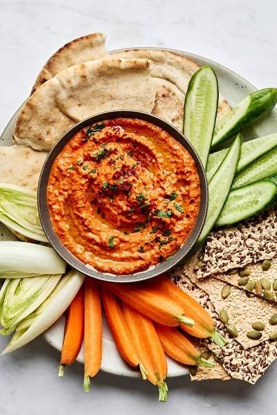 a bowl of homemade Muhammara dip surrounded by crackers, carrots, cucumber and pita for dipping
