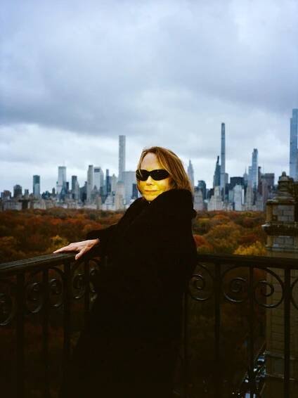 A woman in dark sunglasses and a black coat stands at the corner of a balcony overlooking autumn trees and the New York skyline in the distance.