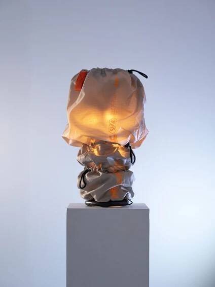 A unique lamp covered in a translucent, drawstring fabric emits a warm glow, displayed atop a simple white pedestal against a soft backdrop.
