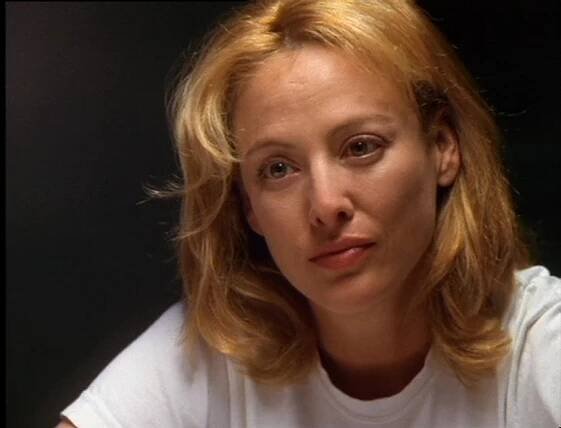 Virginia Madsen as Brenda Kniffen in Just Ask My Children. She sits at a table wearing a plain white T-shirt, her blonde hair slightly tousled. Her expression is weary yet resolute, with a hint of sadness in her eyes. The dim lighting and dark background emphasize the somber and emotional weight of the scene.