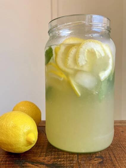 A large glass jar of lemonade with ice, lemon slices and mint. The jar sits on a wooden tabletop next to two fresh lemons..