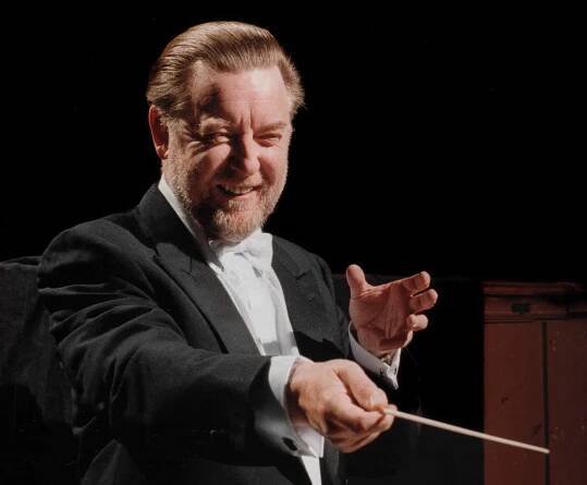 The conductor Andrew Davis in mid-performance, wearing white tie. He has sandy-colored hair and a beard and is gesturing and smiling.