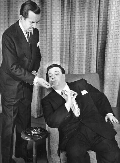 Edward R. Murrow (standing) lighting Gleason's cigarette; Gleason is seated in an armchair