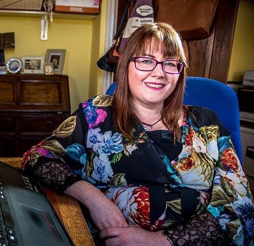 Medium shot of Rebecca Roberts. Rebecca is sitting at her desk at home in a yellow room. She's wearing a black dress with a colourful flower print, rectangle dark red glasses and has medium-length brown hair.
