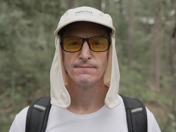 A mean wearing glasses and a hat wearing a backpack with forest in the background.