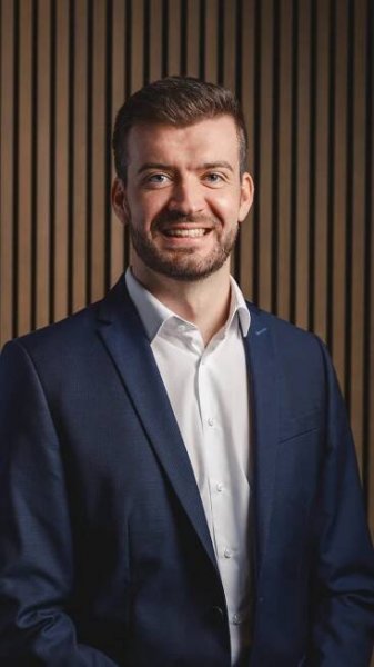 Porträt von Michael Müller, Managing Director Aviation. Er trägt ein dunkelblaues Sakko und ein weißes Hemd. Der Hintergrund besteht aus vertikalen Holzpaneelen, die eine warme und professionelle Atmosphäre schaffen