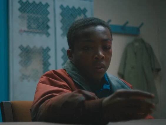 A person in a red jacket looks intently at something in front of them inside a room with patterned walls and a coat on a hook