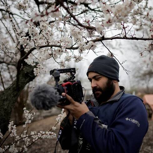 A man filming against a backdrop of a tree with white flowers.