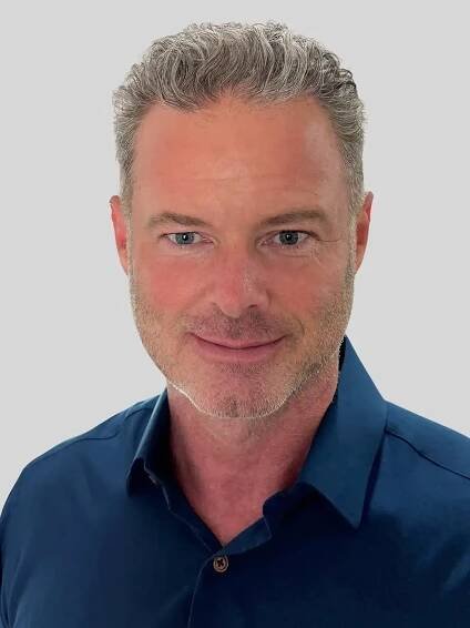 A headshot of a man smiling with silver hair wearing a dark blue button-up shirt