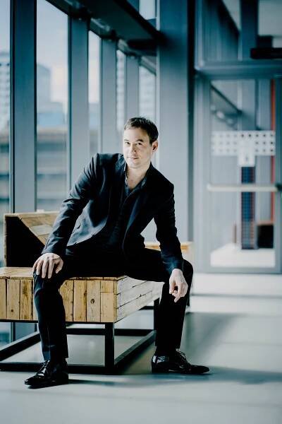 A white man on a wooden seat in front of large windows