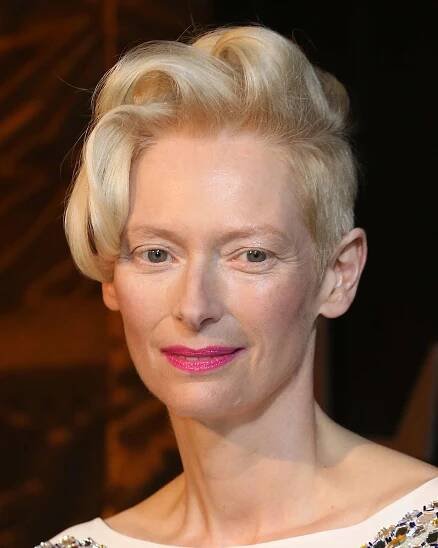 Tilda Swinton wears curly pixie cut, a white top with reflective embroidery, and pink lipstick
