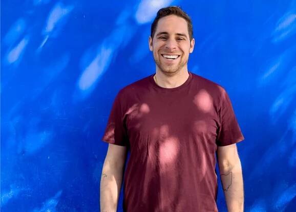 Jonathan Miller smiles in front of a wall painted solid-blue, spotted with sun breaking through shade