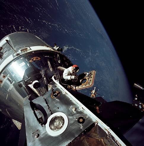 Earth is a deep blue backdrop to this photo of astronaut David Scott and the command module and lunar module of the Apollo 9 mission. The joined modules are a metallic silver. He stands in the open hatch of the command module while wearing a white spacesuit. A metallic visor tops his red helmet.