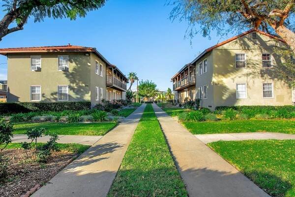 A symmetrical view of a green, landscaped pathway flanked by two rows of apartment buildings. The buildings have small balconies and are surrounded by well-maintained lawns and trees under a clear blue sky, creating a welcoming residential atmosphere.