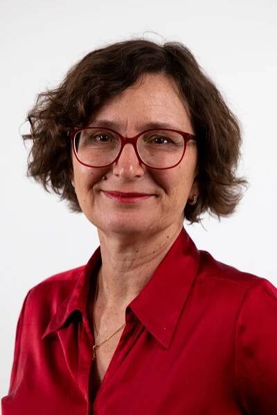 Woman with curly brown hair, wearing red glasses and a red blouse, smiling against a white background.