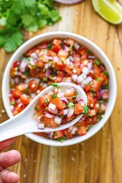 Closet up of pico de gallo on white spoon with bowl of pico de gallo out of focus in the background. 