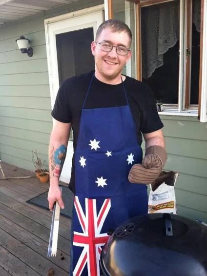 A man is standing next to a webber barbecue. Is wearing glases, a black shirt and an Apron with the Australian flag