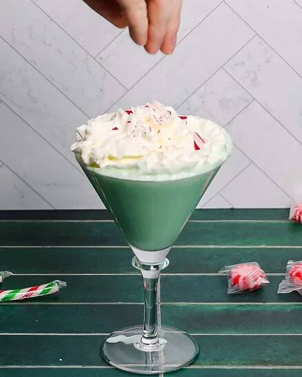 A hand sprinkles crushed peppermint candy over a festive green peppermint martini mocktail, crowned with whipped cream. The drink is elegantly served in a martini glass on a green table, with wrapped peppermint candies nearby, capturing the essence of a delightful Christmas indulgence.