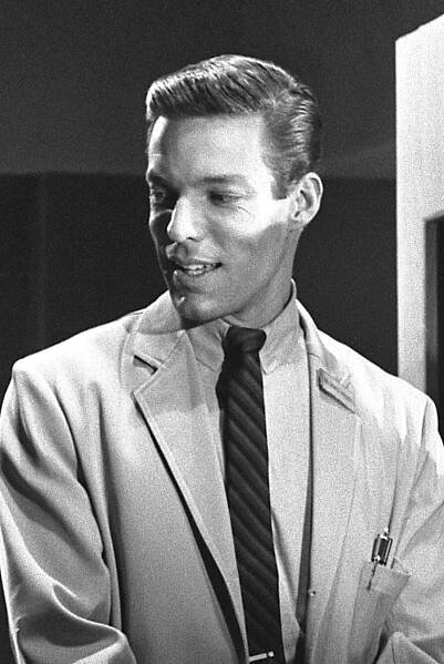 Das schwarz-weiß-Foto von Richard Chamberlain, 1964 trägt er einen Anzug mit Krawatte. Der Anzug hat eine Brusttasche, in der einige Stifte sichtbar sind. Die Szene scheint sich in einem Innenraum abzuspielen. Im Hintergrund sieht man eine dunkle Wand sowie eine hellere Tür oder ein Paneel auf der rechten Seite.