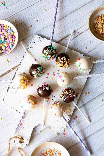 Das Bild zeigt eine Draufsicht auf neun appetitliche Cake Pops, die auf einem rustikalen weißen Holzbrett ausgelegt sind. Sie sind mit Kuchenglasur in Zartbitter und Vanille überzogen, was eine schöne Farbvielfalt erzeugt. Einige der Kuchenkugeln am Stiel sind mit gehackten Nüssen oder Krokant dekoriert, während andere mit einer bunten Mischung aus Streuseln und bunten Zuckerperlen verziert sind. Einige der Cake-Pop-Stiele sind weiß, andere lila-weiß gestreift, was dem Bild eine spielerische Note verleiht. Im oberen linken Bereich des Bildes ist eine kleine Schüssel mit weiteren farbenfrohen Streuseln zu sehen, während oben rechts eine Schüssel mit gehackten Nüssen oder Krokant platziert ist, bereit für die Dekoration. Einzelne verstreute Streusel liegen auf dem hellen Holzhintergrund und verstärken den Eindruck eines kreativen Backprozesses für diese einfachen und beliebten Leckereien, die perfekt für kleine Kinderhände sind und leicht mit dem Thermomix® zubereitet werden können.