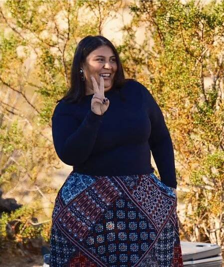 A woman smiling and standing outdoors, wearing a dark top and a colorful patterned skirt. She is making a peace sign with her right hand against a backdrop of greenery.