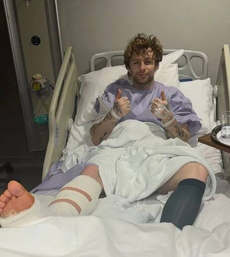 Tom Grennan in a hospital bed with a leg injury, giving a thumbs-up.