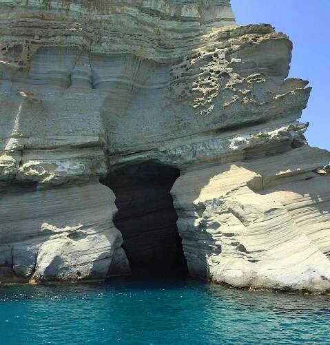 A rock formation with a cave situated in water, surrounded by cliffs and natural arch features. The scene showcases the beauty of erosion in nature, highlighting a serene body of water adjacent to the rugged landscape.
