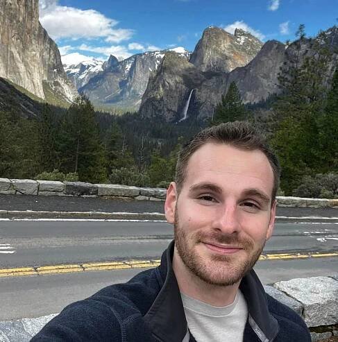 A man is taking a selfie in front of a mountain range.