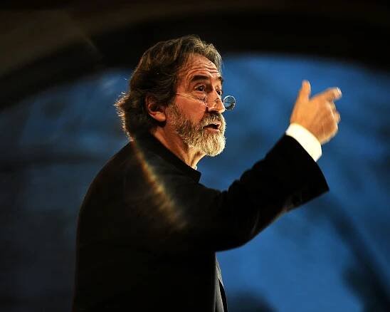 Jordi Savall mit grauem Haar und Bart gestikuliert leidenschaftlich. Er trägt eine Brille und einen schwarzen Anzug. Er tritt vor einem unscharfen Hintergrund auf, der von dramatischer Beleuchtung angestrahlt wird, die einen Regenbogeneffekt erzeugt.