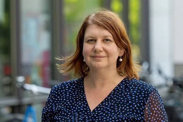 Dieses Bild zeigt Prof. Dr. Yulia Golub, eine Frau mit rötlichem Bob und Perlenohringen. Sie trägt eine schwarze Bluse mit blauem Tupfenmuster und schaut freundlich in die Kamera. Im Hintergrund sind verschwommen ein Gebäude und parkende Fahrräder zu sehen.