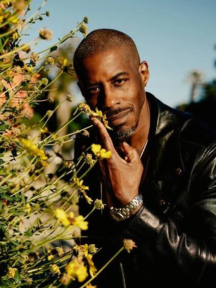 A man with a goatee and mustache leans toward a tall plant and pulls a flower toward his face. He’s smiling softly and is wearing a black leather jacket and a silver watch.