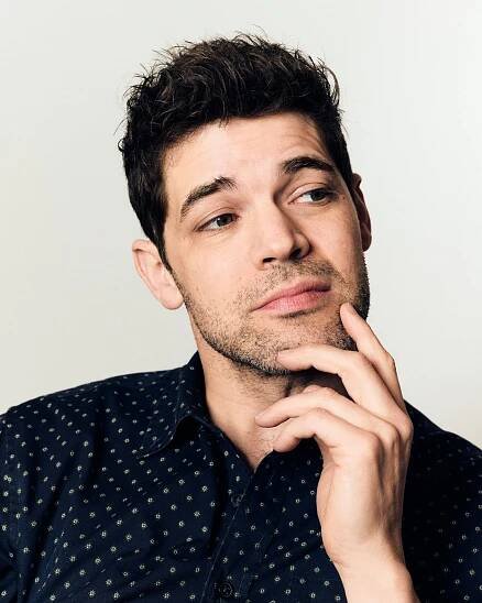 A young man looks off to the side, the fingers of his left stroking his beard.