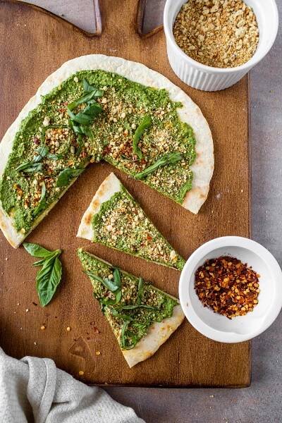 vegan pesto pizza on wood platter with red pepper flakes and parmesan cheese