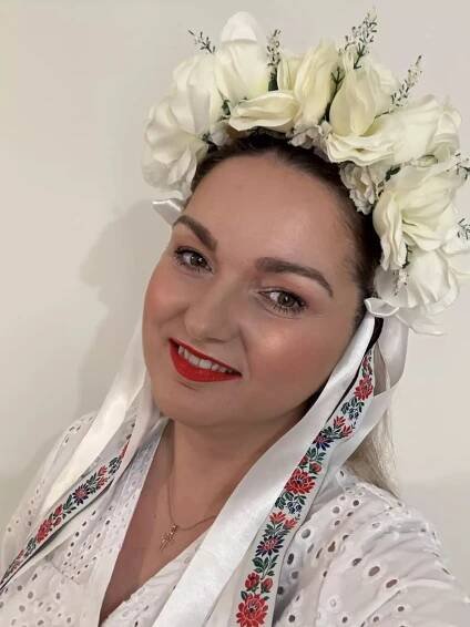Usmievavá žena v bielej kvetinovej čelenke a tradičnom vyšívanom odeve s červenými a modrými vzormi, vyjadrujúca radosť a kultúrnu hrdosť.