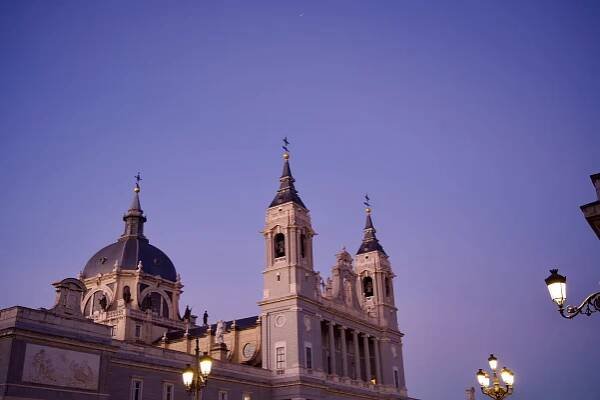 The Almudena Cathedral in Madrid at dusk, its white Baroque facade illuminated by street lamps against a purple sky