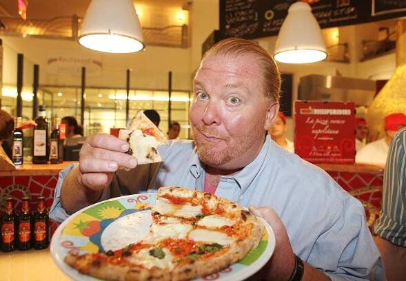 Mario Batali holding up a slice of Pizza at a restaurant.