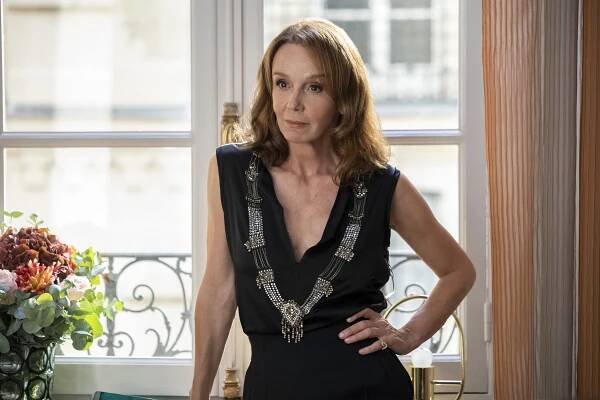 A woman in a sleeveless v-neck black dress with a statement necklace, one hand on her hip implying attitude as she stands in front of a set of French doors; still from "Emily in Paris"