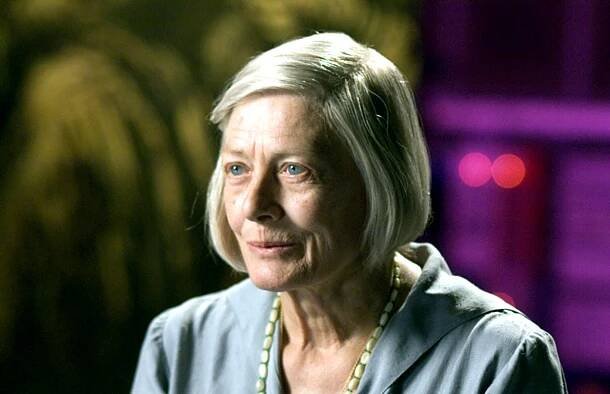 Person with short hair wearing a gray collared shirt and a necklace, set against a blurred backdrop
