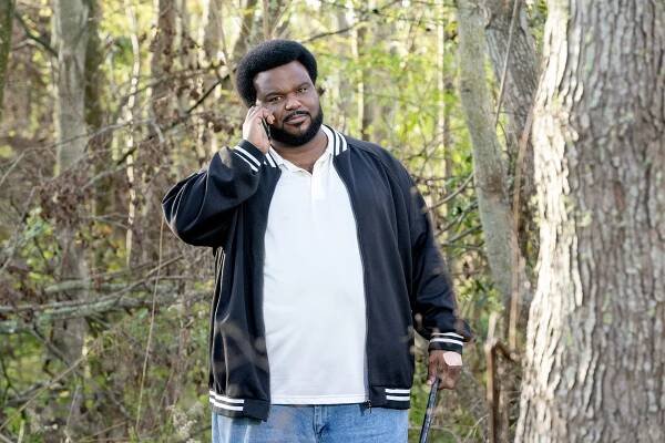 Craig Robinson stands in the woods talking on a cell phone.