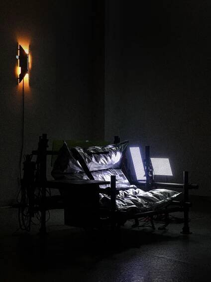 A dimly lit room featuring an industrial-style bed with metallic pillows, flanked by two bright lights, creating a stark contrast.