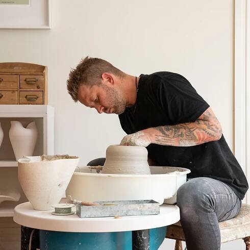 Man shaping clay on a pottery wheel in a studio, focused and surrounded by pottery tools and materials.
