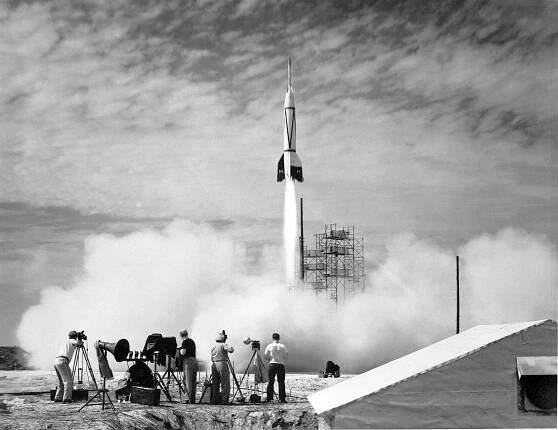 In the foreground, four people stand outside near equipment as a white rocket launches in front of them. White vapor clouds cover the ground and much of the scaffolding at the launch site. The photo is black and white.